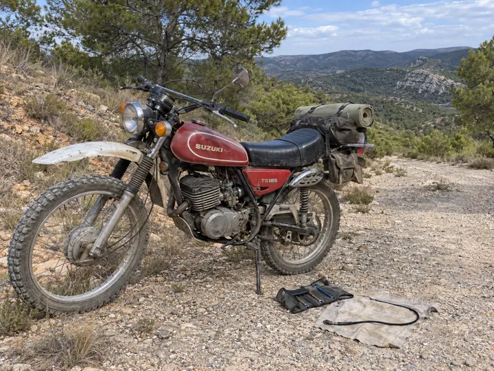 A weathered red Suzuki TS185 with camping luggage, tools and a detached cable beside a dusty trail in Spain.